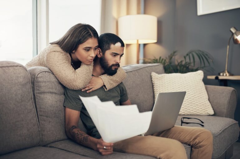 A man leans over a woman's shoulder looking at an open laptop. The woman is sitting at a wooden table.