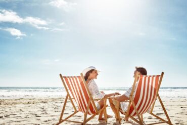 A smiling couple holds hands while sitting in orange striped beach chairs on the sand near the ocean.