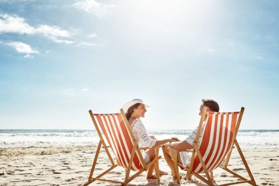 A smiling couple holds hands while sitting in orange striped beach chairs on the sand near the ocean.