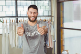 A bearded man looks toward the camera and smiles. He holds two thumbs up and wears a shirt that says "Volunteer."