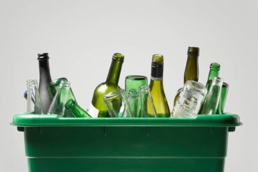 A green, plastic bin contains several empty glass bottles. There are green, clear, and black bottles.