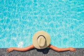 A person wearing a sun hat stretches their arms across the tiled edge of a swimming pool as they recline against it.