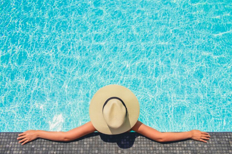 A person wearing a sun hat stretches their arms across the tiled edge of a swimming pool as they recline against it.