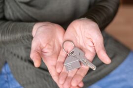 A person wearing a gray shirt and blue bottoms holds a keychain with a silver house and two keys on it in their palms.