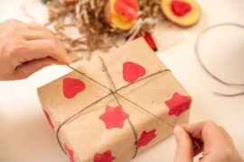 A person ties a light string into a knot around a small box wrapped in handmade paper with a red printed pattern.