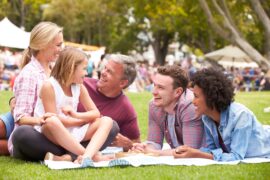 A diverse group of people relaxing together in the grass. Other people under event tents are visible in the background.