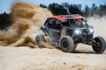 A red and black UTV buggy with a driver and passenger in the desert driving at fast speeds and kicking up clouds of sand.