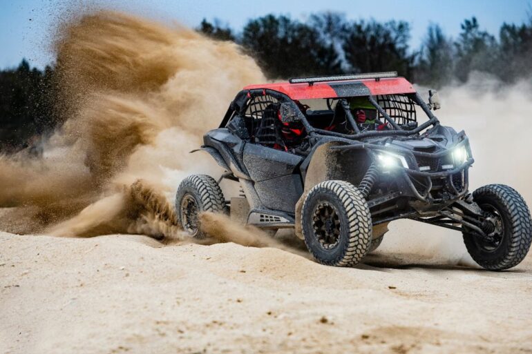 A red and black UTV buggy with a driver and passenger in the desert driving at fast speeds and kicking up clouds of sand.