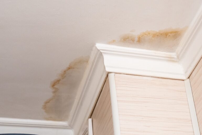 Close up of a white ceiling, white crown molding, and light tan walls. The ceiling has an orange-brown water stain.