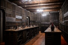 A wine storage room with dark wood shelving units of wine bottles and a counter with wine glasses on it inside.