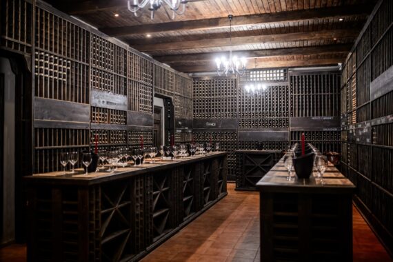 A wine storage room with dark wood shelving units of wine bottles and a counter with wine glasses on it inside.