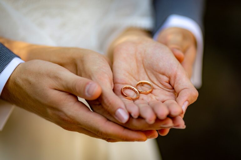A couple in formal attire with their hands cupped together holds two golden wedding band rings outside.