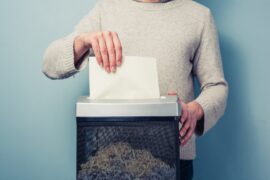 A close-up view shows a person placing a piece of blank, white paper into a black and gray paper shredder.