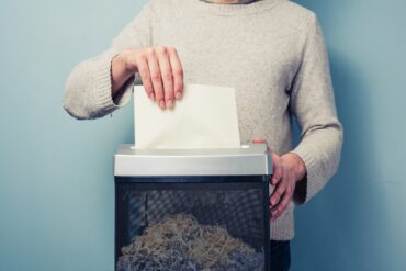 A close-up view shows a person placing a piece of blank, white paper into a black and gray paper shredder.