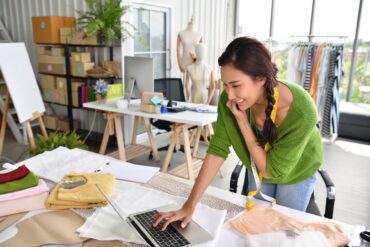 A woman wearing a green sweater, with measuring tape around her neck, uses a laptop in a room filled with clothes and boxes.