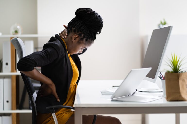 A woman wearing black and yellow clothes sits at a desk while touching her neck and back as if she's in pain.
