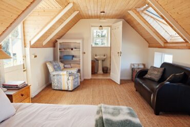 A cozy attic bedroom with a couch, a corner chair, a bed, and a small bathroom, under slanted ceilings.