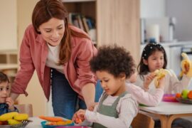 An older woman wearing pink long sleeves and jeans leans over a group of younger children eating fruits at a table.