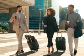 Three well-dressed professionals walking and talking; they're carrying bags, suitcases, and coffee on their way to work.