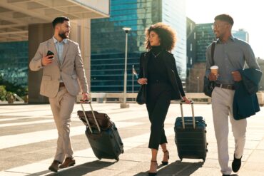 Three well-dressed professionals walking and talking; they're carrying bags, suitcases, and coffee on their way to work.