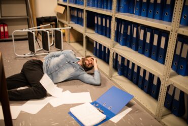 A man lying on the floor, surrounded by papers, appearing to be wincing in pain, next to a step ladder he possibly fell from.