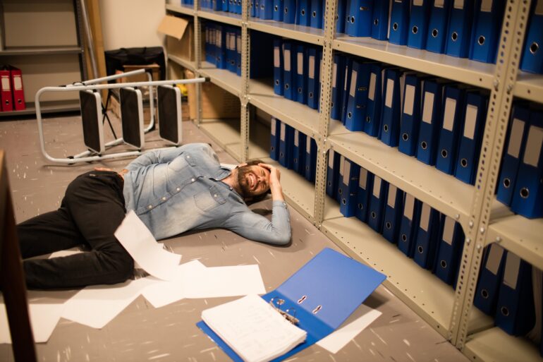 A man lying on the floor, surrounded by papers, appearing to be wincing in pain, next to a step ladder he possibly fell from.