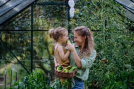 A woman wearing a green shirt and blue jeans holds a young child and a basket of vegetables while standing in a greenhouse.