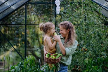 A woman wearing a green shirt and blue jeans holds a young child and a basket of vegetables while standing in a greenhouse.