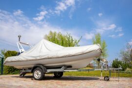 A boat covered with a white tarp sits on a boat trailer on a brick driveway. Trees and a blue sky appear behind it.