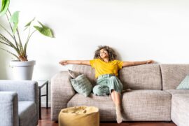 A Black woman is happily relaxing on the grey/beige sofa at home. The couch has light teal throw pillows and a yellow pouf.