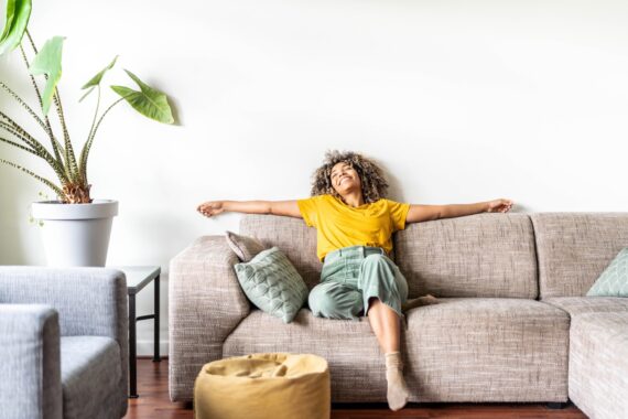 A Black woman is happily relaxing on the grey/beige sofa at home. The couch has light teal throw pillows and a yellow pouf.