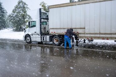 A white semitruck pulled over to the side of a snowy highway. The driver is inspecting the side of the trailer.