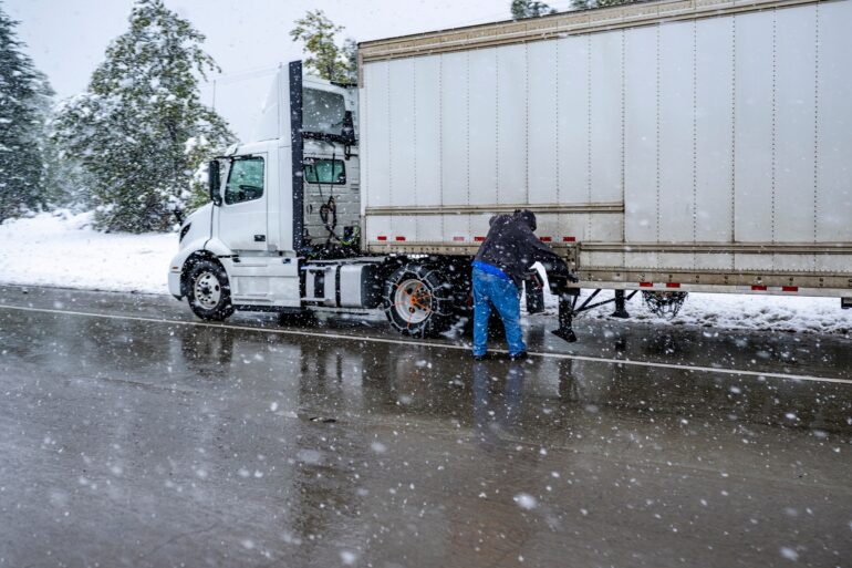 A white semitruck pulled over to the side of a snowy highway. The driver is inspecting the side of the trailer.