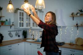 A smiling young woman in a red and black flannel screws a lightbulb into a light fixture in a kitchen.
