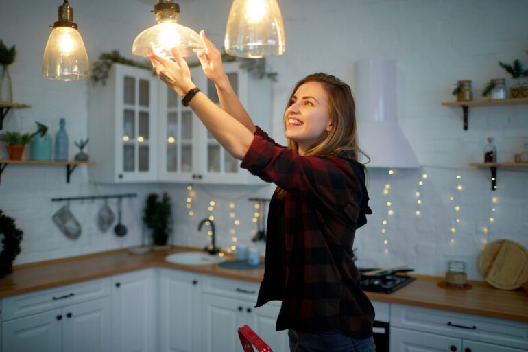 A smiling young woman in a red and black flannel screws a lightbulb into a light fixture in a kitchen.