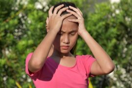 A girl in a pink shirt holds her head with both hands, standing outdoors in front of a softly blurred plant.