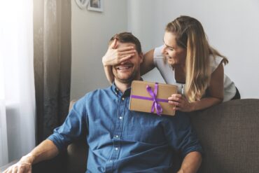 A man sitting on a couch while a woman covers his eyes from behind and leans over his shoulder to give him a wrapped present.