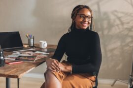 A young Black woman sitting at a desk in a simple office space. The desk has a laptop and several photos sitting on it.