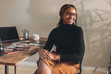 A young Black woman sitting at a desk in a simple office space. The desk has a laptop and several photos sitting on it.
