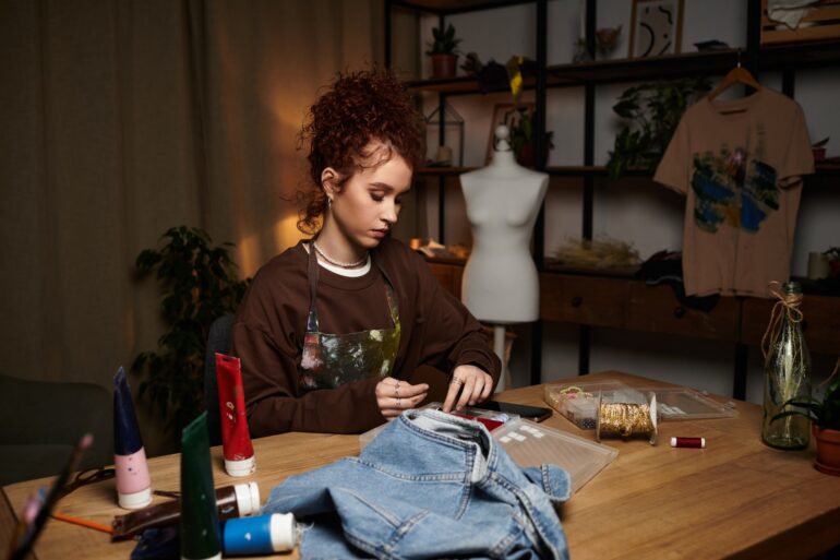 A woman wearing an apron sits at a wooden table with art supplies, paint tubes, and a denim vest laid out on it.