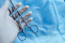 A medical professional wears a white latex glove while holding a pair of stainless steel lab scissors.