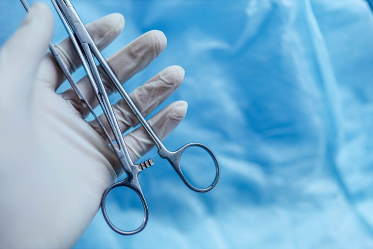 A medical professional wears a white latex glove while holding a pair of stainless steel lab scissors.