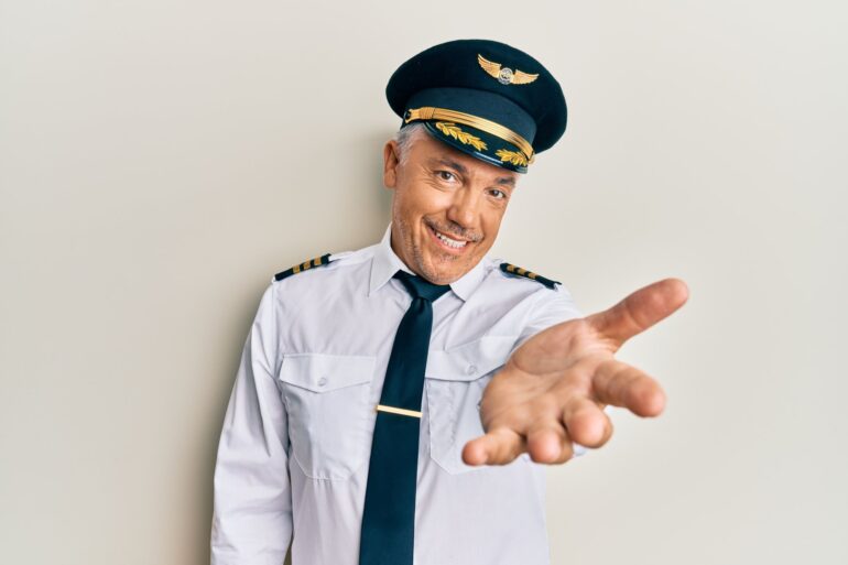 An older gentleman, dressed in a white shirt and blue pants as his pilot uniform, offers a welcoming palm while smiling.