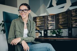 A young woman smiles as she sits in a black office chair. The wooden desk behind her contains two monitors.