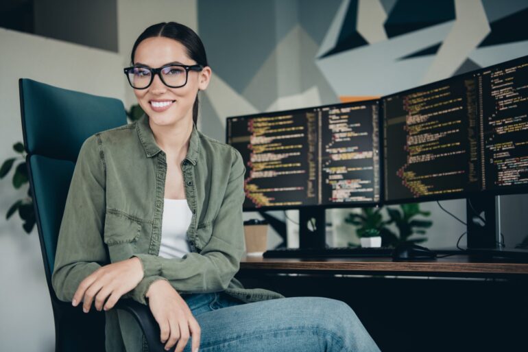 A young woman smiles as she sits in a black office chair. The wooden desk behind her contains two monitors.