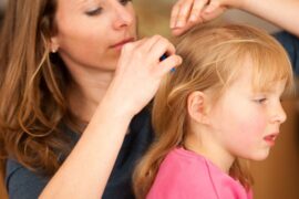 A woman wearing a blue shirt sits next to a little girl wearing pink while combing through her hair for lice.