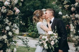 A bride and a groom are kissing, surrounded by various white flowers and green leaves, while standing outside.