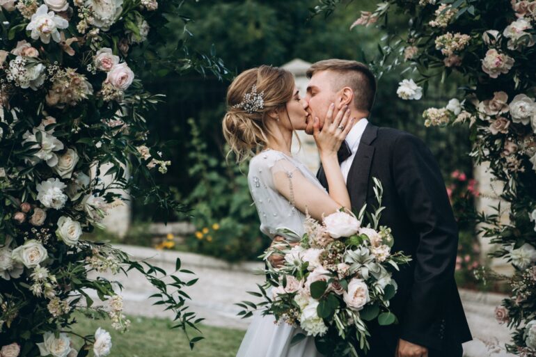 A bride and a groom are kissing, surrounded by various white flowers and green leaves, while standing outside.