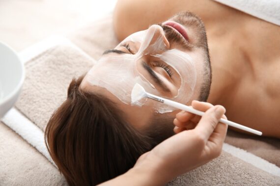 A bearded man lying on a towel with an aesthetician applying a mask to his face with a small brush.