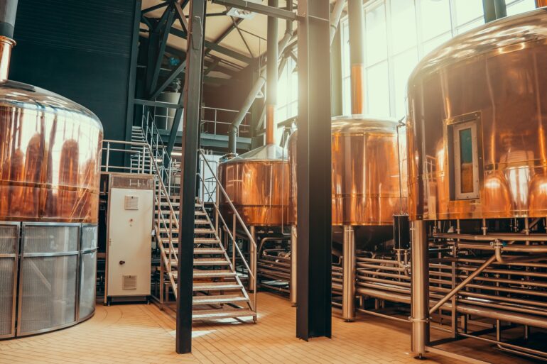 A brewery operations room with large brew tanks and access stairwells. The large tanks are copper colored.